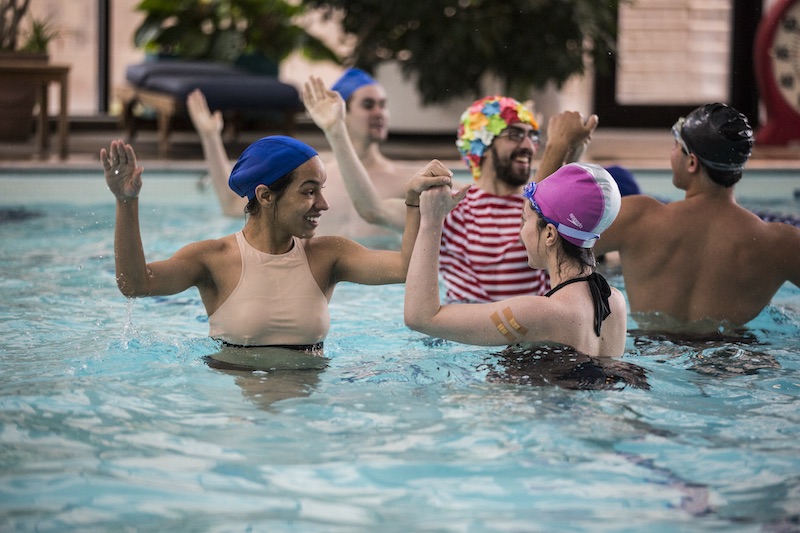 Performers in bath caps and suits pair off in the pool. They reach clasp their partners hand as their stand in the shallow end.